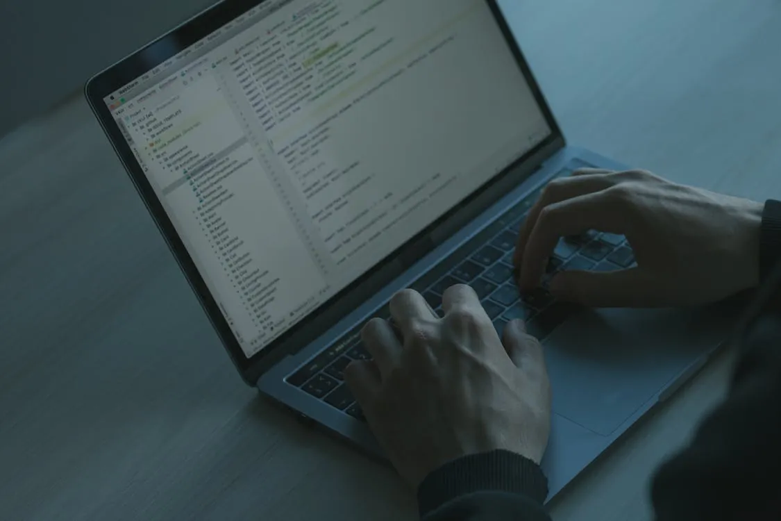 The photo shows a person working on a laptop with code or text editor open, situated on a light-colored surface, highlighting programming or technical work in a dimly lit environment, suitable for articles about software development or remote work.