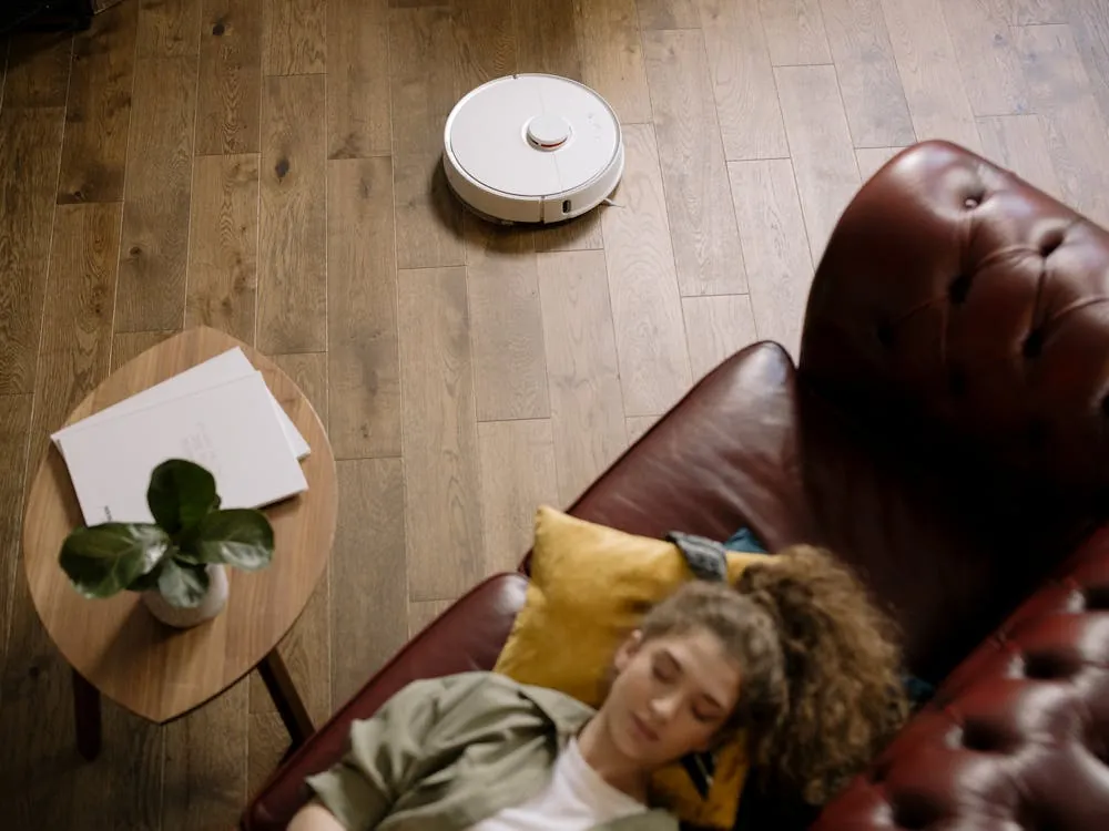 The image shows a woman resting on a brown leather sofa with a yellow pillow, lying underneath a child's blanket, in a cozy living room with wooden flooring. A robotic vacuum cleaner is cleaning the floor nearby, while a small wooden side table holds a potted plant and some papers.