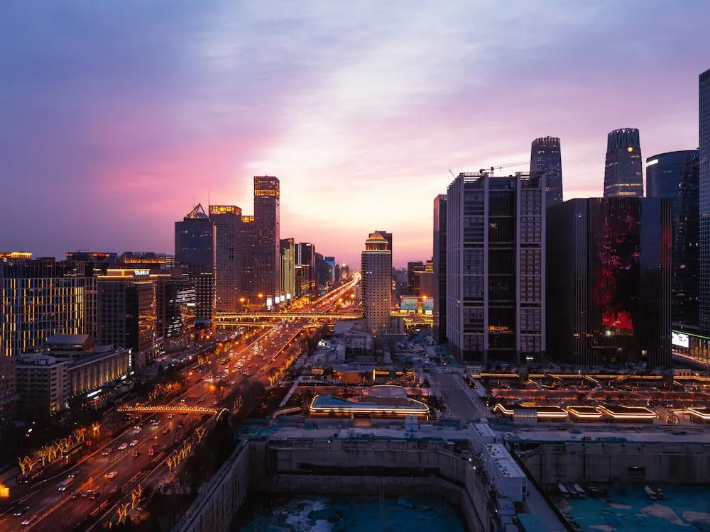 This photo shows a bustling modern city skyline at sunset, featuring tall skyscrapers with illuminated windows and busy streets filled with moving traffic. The vibrant sky shifts from purple to pink, highlighting the urban environment and dynamic atmosphere suitable for topics related to city life, architecture, or evening cityscapes.