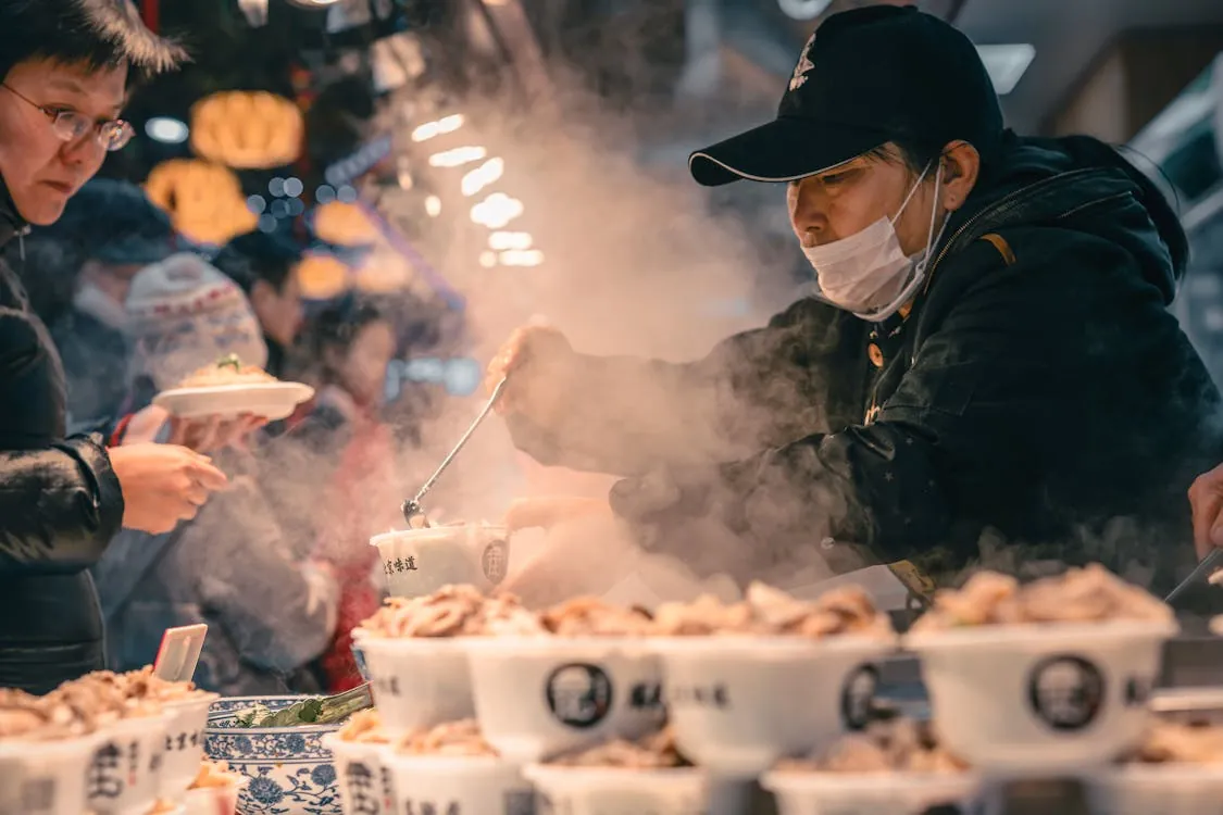 Bustling street food scene in Beijing with vendor serving steamed dishes to customers in the night market.