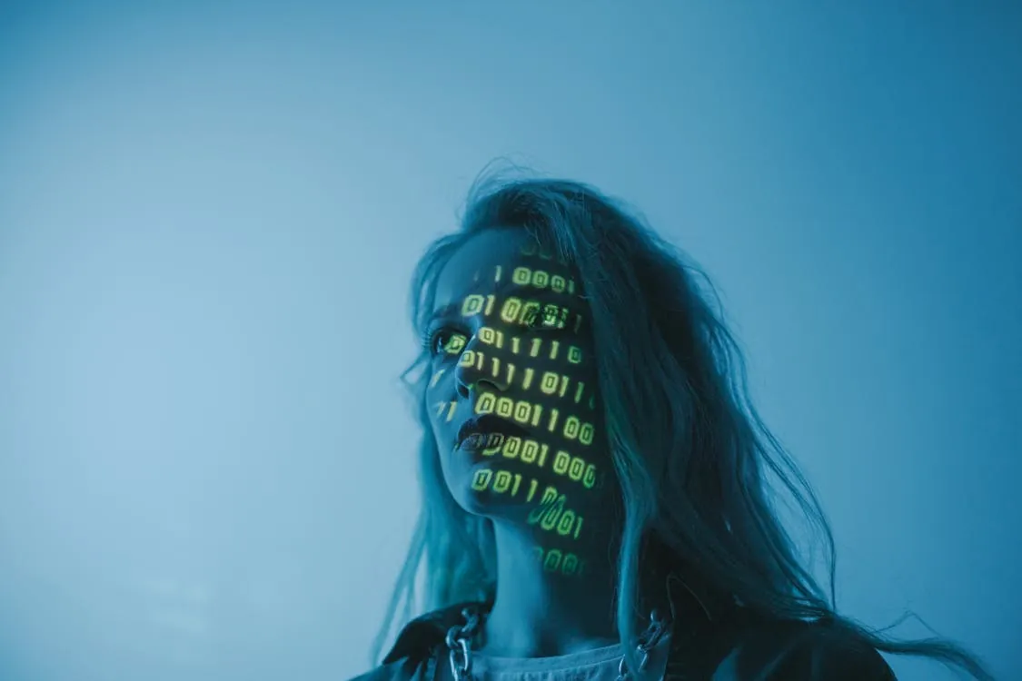 This photo features a woman with long hair illuminated by green digital binary code projected onto her face, set against a cool blue background, capturing a futuristic and cyberpunk aesthetic. It would suit articles on technology, digital innovation, or cybersecurity themes.