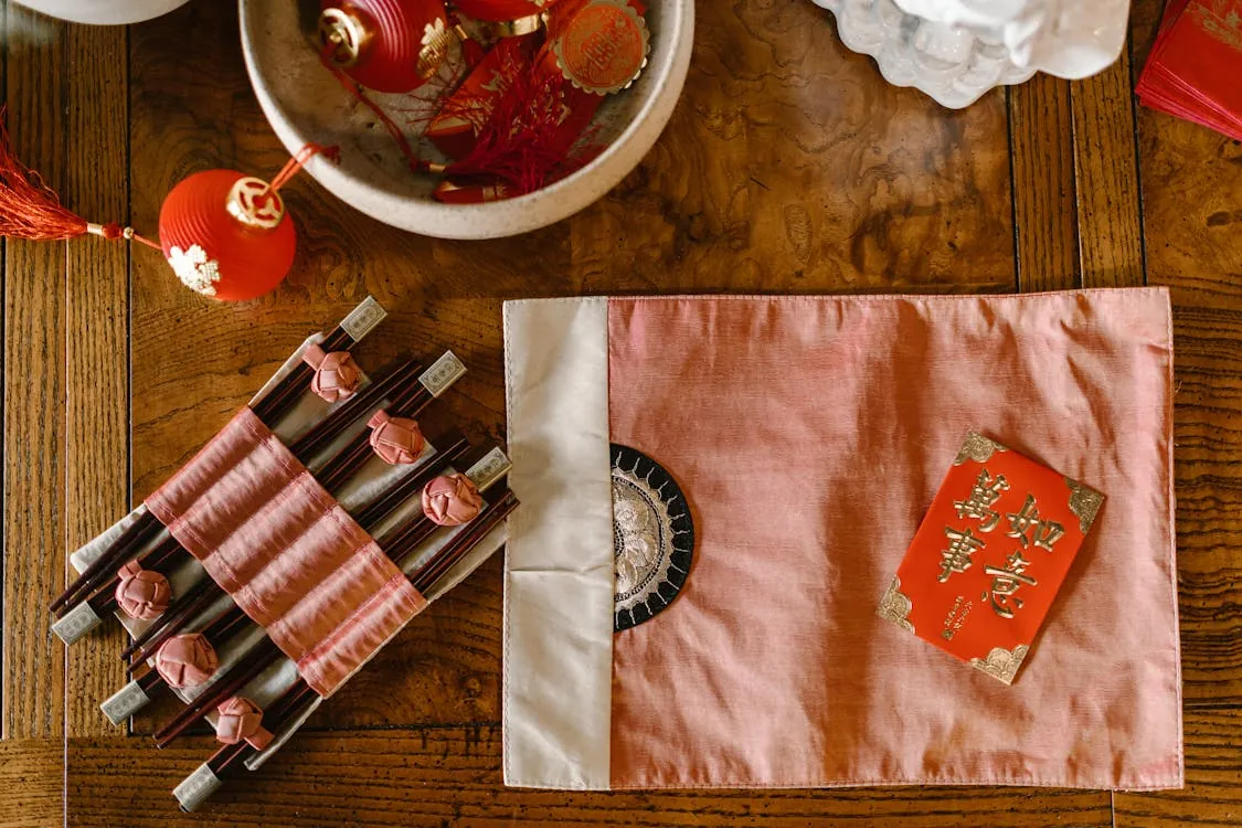 This image features a festive Chinese New Year setup on a wooden table, including red and gold decorations, a bowl with ornaments, wrapped chopsticks with pink embellishments, and a pink satin cloth with a red envelope on top. The scene highlights traditional celebration elements, making it suitable for articles about Chinese holidays, cultural customs, or New Year festivities.
