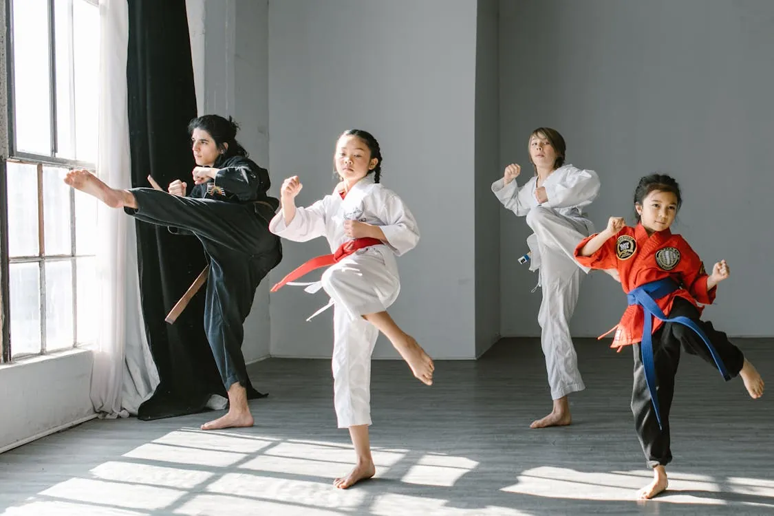The photo shows four children practicing martial arts in a well-lit indoor studio, with three wearing traditional gi uniforms and one in a red martial arts gi, all performing high kicks and stances on a wooden floor. The setting emphasizes a disciplined, active training environment suitable for articles about martial arts classes for kids or youth fitness programs.