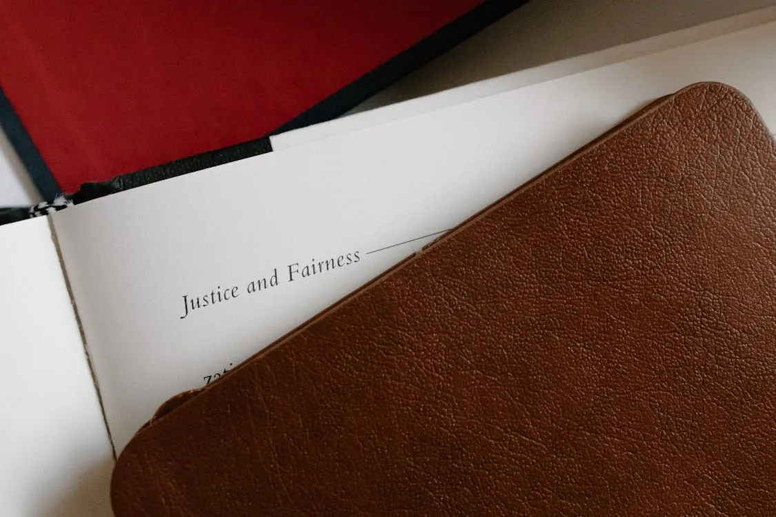 This photo shows a close-up of an open book or booklet with the title "Justice and Fairness" visible on the page, partially covered by a brown leather cover or folder, set against a background that includes a red object or surface. It suggests a legal, ethical, or academic context, suitable for an article discussing justice, fairness, or legal principles.
