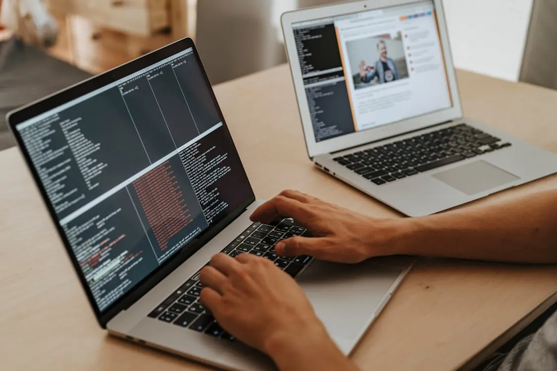 This photo shows a person working on laptops, with one displaying lines of code or data in a dark-themed terminal, and the other showing a social media feed or article featuring a person speaking. The setting appears to be a workspace or home office, highlighting themes of programming, digital communication, or online content creation.