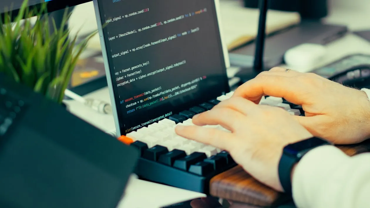 This photo shows a person coding on a laptop with visible programming code on the screen, sitting at a desk that includes a small potted plant, a computer mouse, and a keyboard, suggesting a tech or software development environment. The focus is on the individual's hands typing, making it suitable for articles related to programming, software engineering, or remote work.