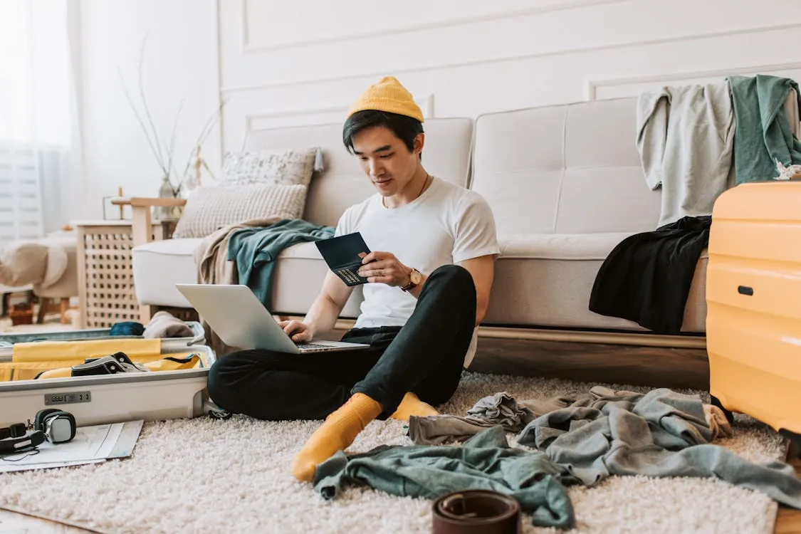 A young man in a messy living room preparing for travel with a laptop and passport, representing digital nomads in Spanish and English speaking countries.