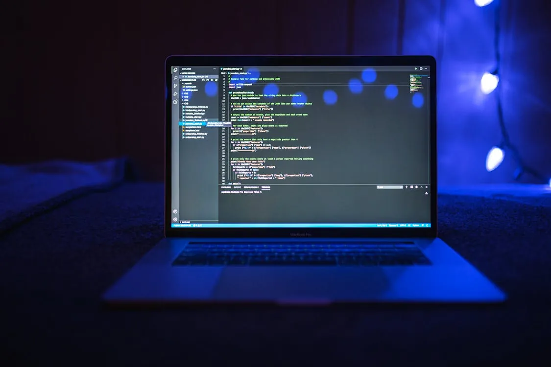 This photo shows a dark room with a laptop displaying code in a text editor, illuminated by the screen and a few bokeh-style blue lights in the background. It highlights a tech or programming environment, suitable for articles about software development, coding, or computer science.
