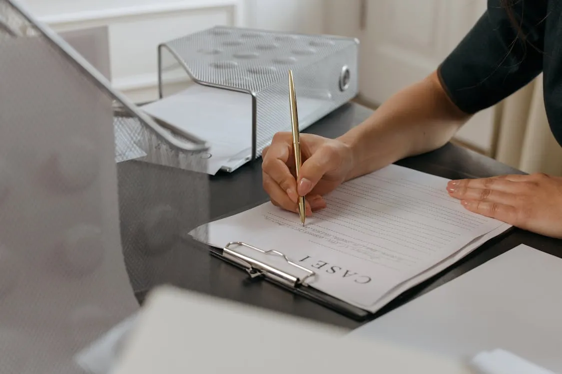 The image shows a person filling out a contract form on a clipboard at a desk, with various office supplies such as a file organizer and papers nearby. The setting appears to be an office or professional environment, suitable for articles about legal agreements, onboarding processes, or administrative tasks.