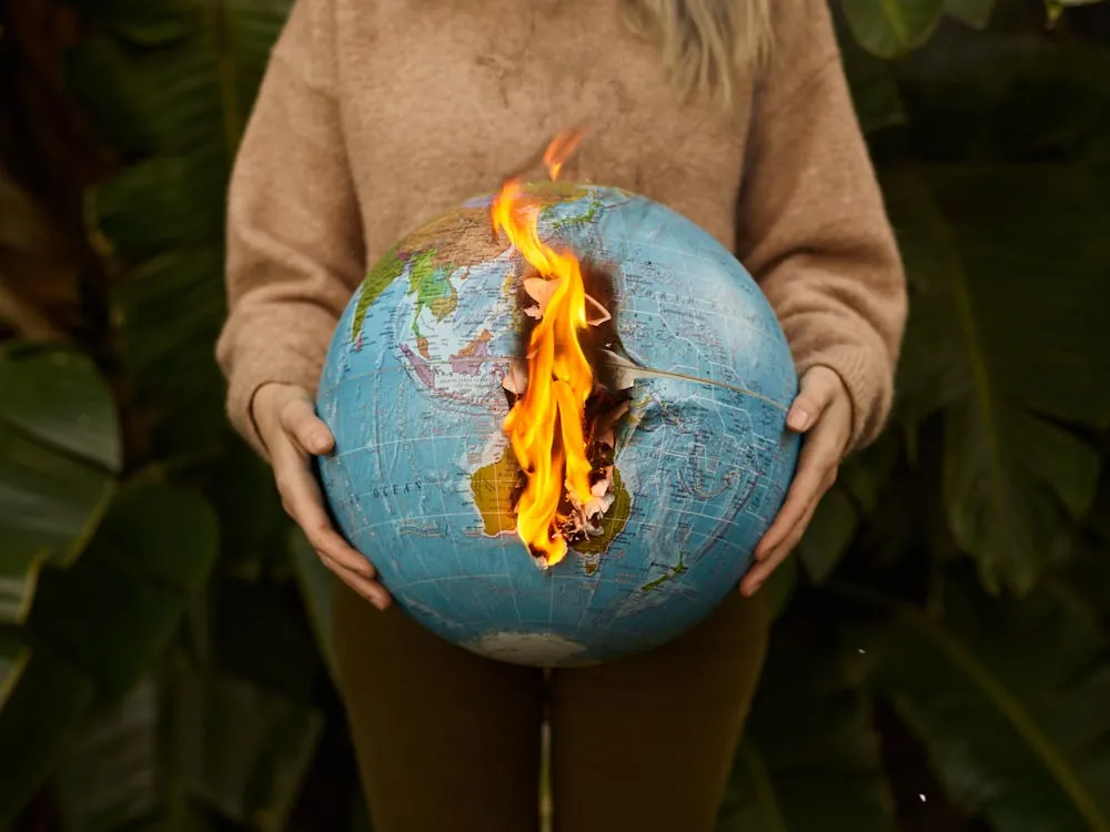 The image shows a person holding a globe with a large, realistic fire burning along a vertical crack, symbolizing environmental destruction or climate change. The setting appears to be outdoors, surrounded by green foliage, and the focus is on the stark contrast between the globe and the flames.