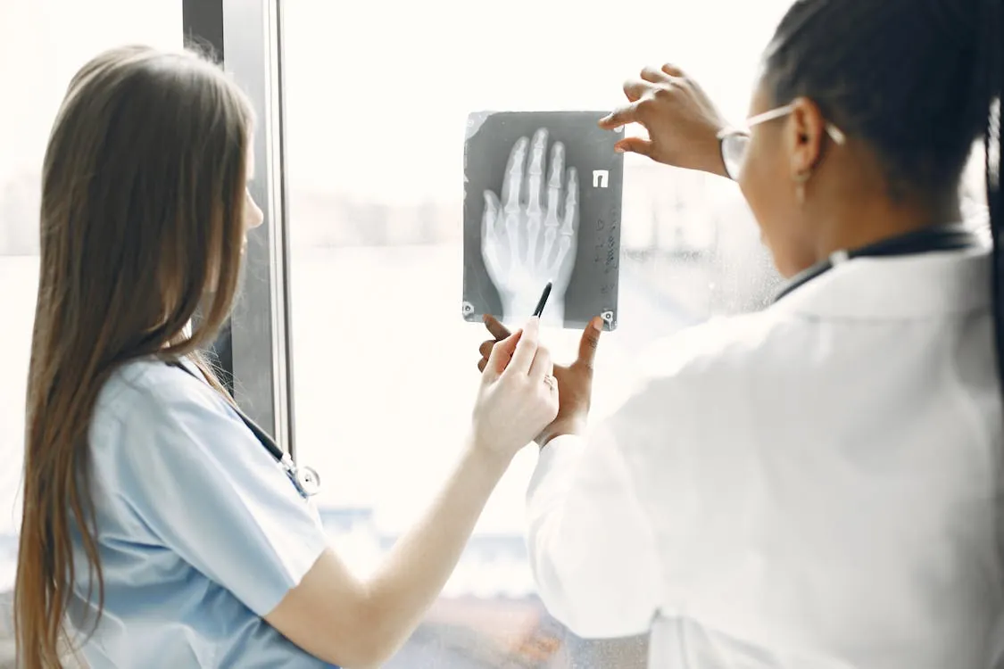 The image shows two medical professionals, likely doctors or radiologists, analyzing and discussing an X-ray image of a human hand near a window, with one holding a pen and pointing at the X-ray. This setting emphasizes a clinical, diagnostic environment suitable for articles on healthcare, medical research, or radiology.