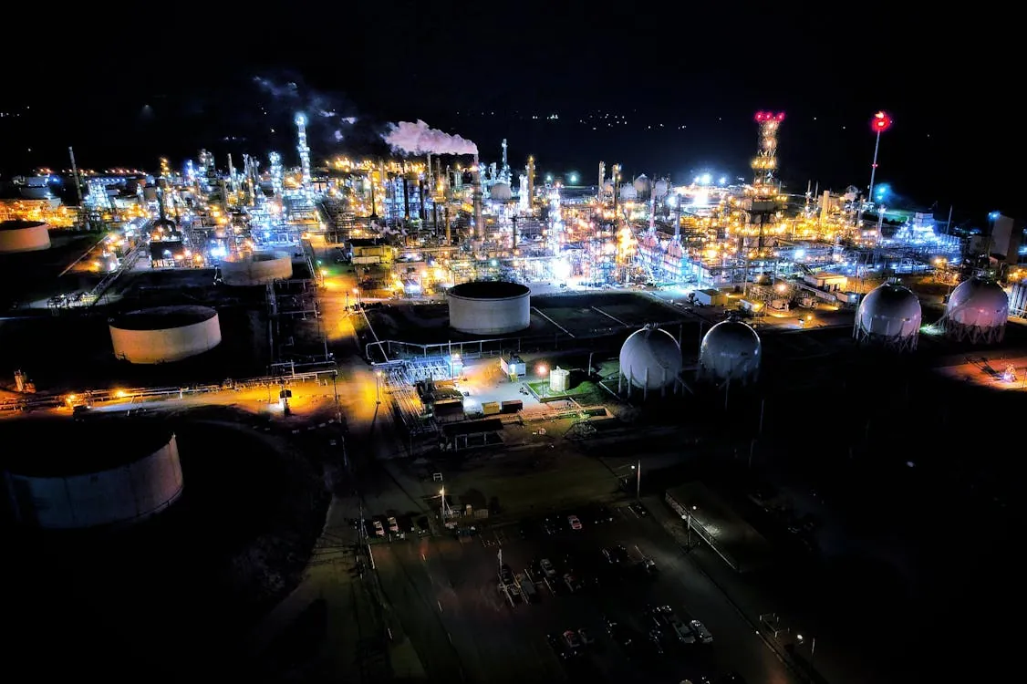 Aerial nighttime view of an illuminated oil refinery complex with tanks and pipelines.