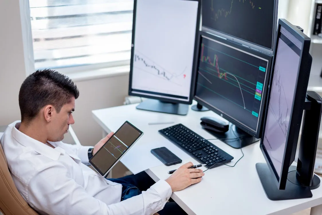 Trader at a desk with multiple monitors showing financial charts and a tablet