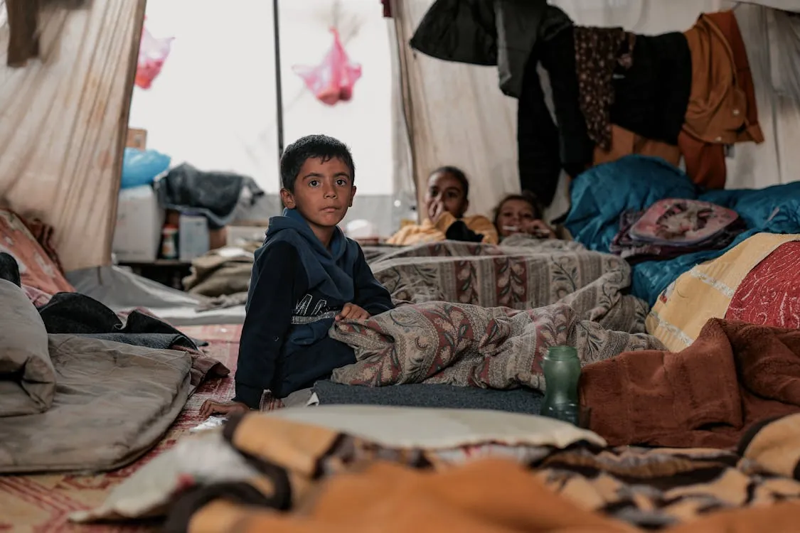 Children sitting in a makeshift shelter, highlighting displacement from war in Iran