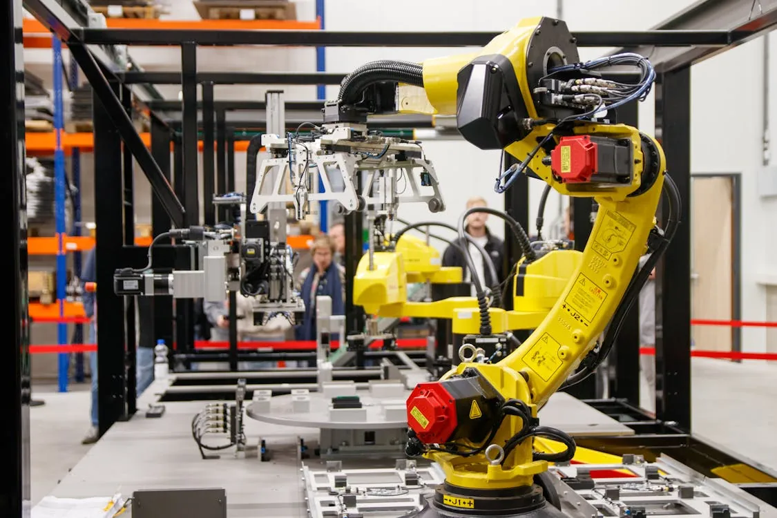 The photo shows a yellow industrial robotic arm working on an assembly line inside a manufacturing facility, with several people observing in the background; it highlights advanced automation technology used for industrial production or assembly processes.