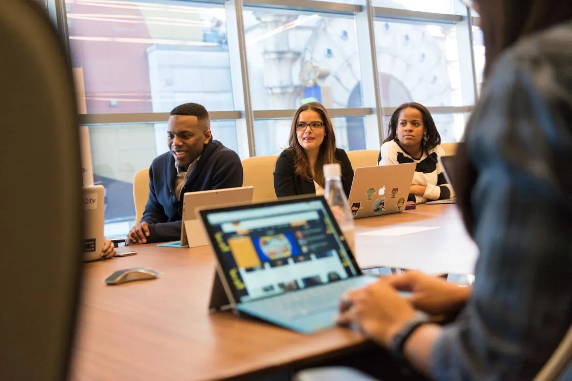 The image shows a diverse group of professionals and colleagues engaged in a meeting or discussion around a conference table in a modern office or coworking space, with large windows behind them providing natural light, and laptops and tablets in use. This setting would suit articles on workplace collaboration, remote work, or tech-driven professional environments.