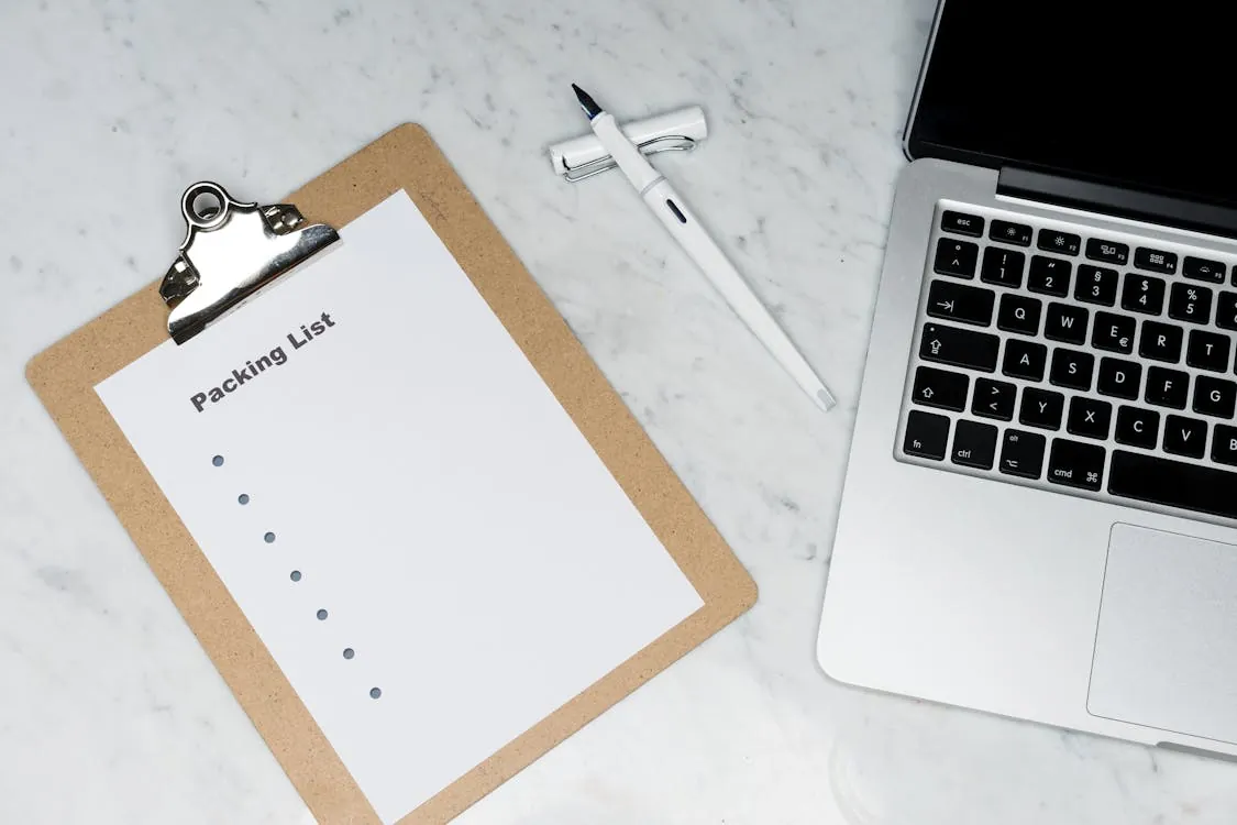 This image shows a top-down view of a workspace featuring a clipboard with a "Packing List" sheet, a white pen, a laptop, and a partially visible smartphone on a marble surface. It is suitable for articles related to travel preparation, organization, or productivity tips.