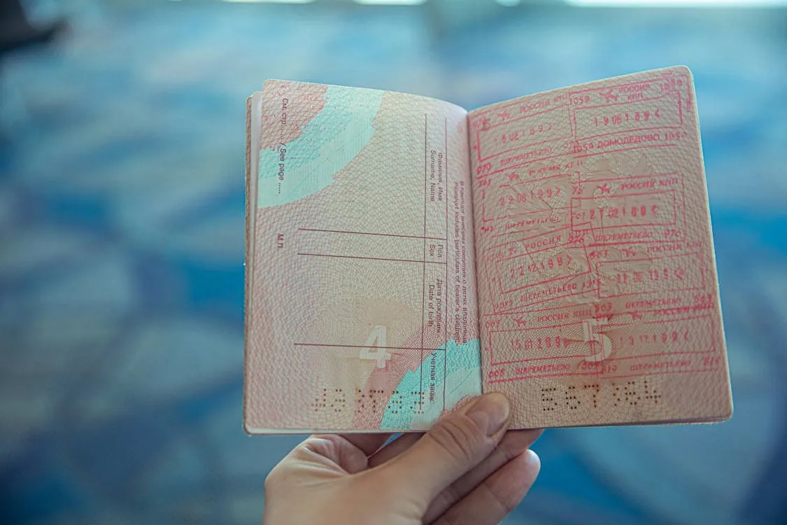 This photo shows an open passport being held in front of a blurred blue background, with multiple visa stamps and entry/exit markings visible on the right page, indicating international travel. The image would suit a blog article related to travel, visas, or the passport application process.