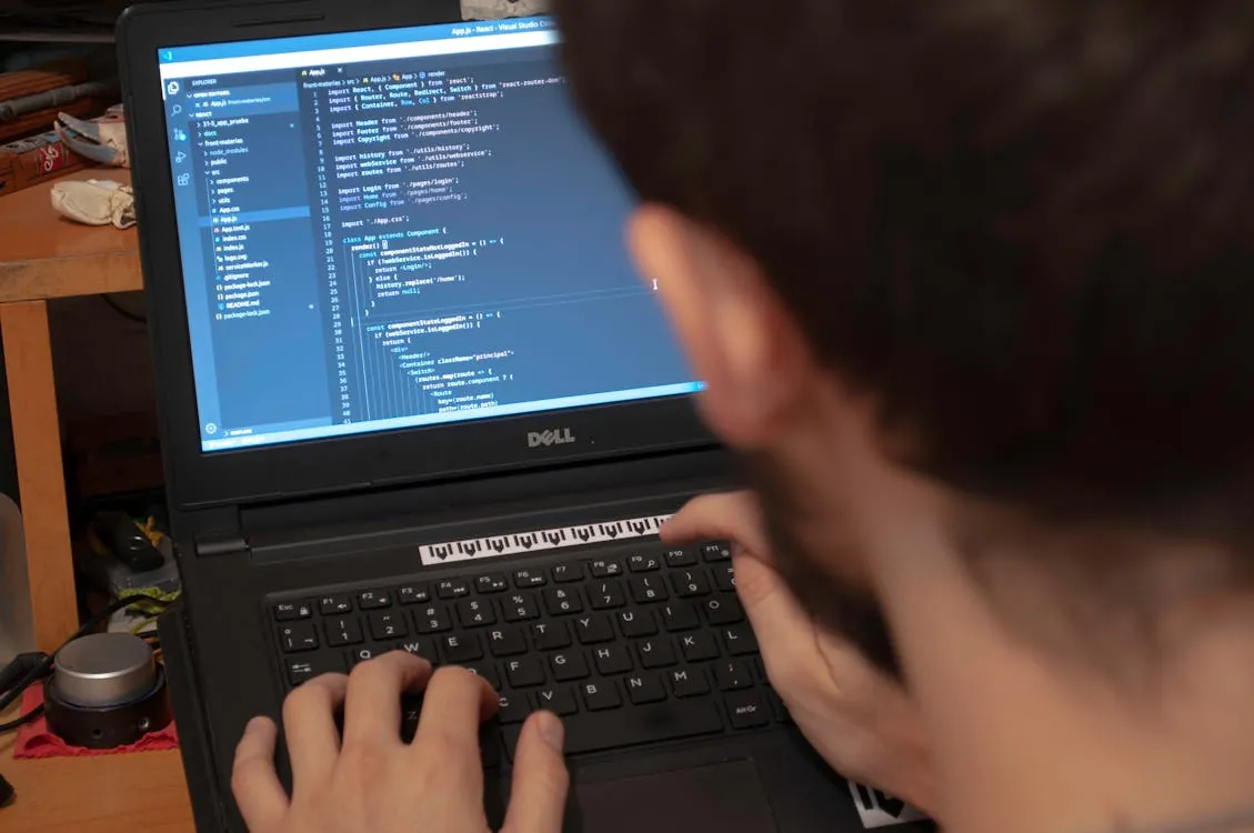The photo shows a person coding or programming on a Dell laptop, with the screen displaying lines of code, likely in a software development environment. The setting appears to be a workspace or desk with some objects in the background, indicating a focus on tech, programming, or software engineering.