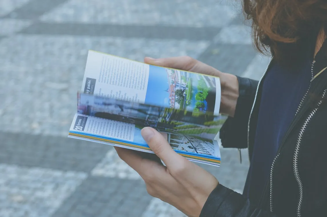 The photo shows a person holding and flipping through a colorful travel or city guidebook outdoors on a paved surface, with a focus on the open pages featuring images and text about urban scenery. This image would be suitable for articles related to travel planning, urban exploration, or sightseeing tips.