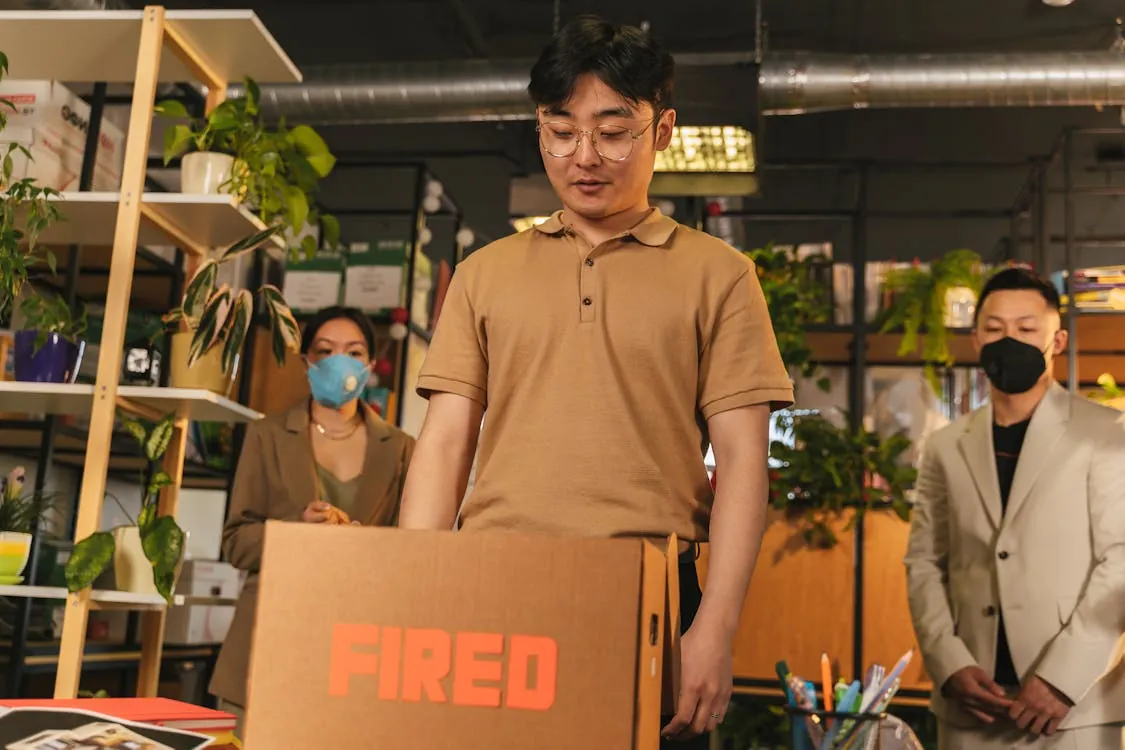 The image shows a young man in a beige polo shirt lifting a cardboard box labeled "FIRED" in an office-like environment, with two colleagues in the background wearing masks—one woman in a brown blazer with a blue mask and a man in a white blazer with a black mask—surrounded by shelves with plants and office supplies, suggesting a workplace setting related to a staff dismissal or office transition.