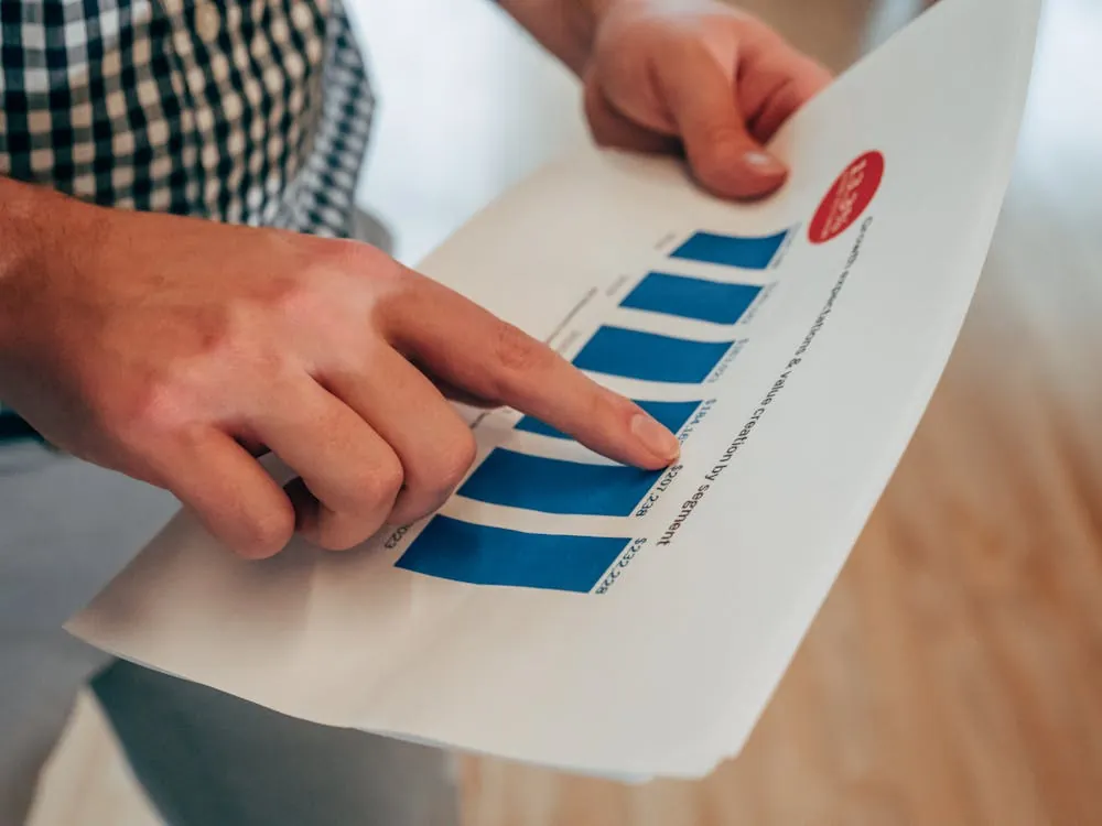 This photo shows a close-up of a person pointing at a printed bar graph on a sheet of paper, likely during a discussion or presentation. The setting appears to be an indoor environment, possibly at a desk or conference table, with the focus on data analysis or work-related activities.