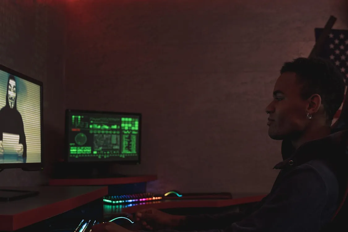 The photo shows a young man sitting in a dimly lit room, working on a computer with multiple monitors displaying hacking-related visuals and green code, suggesting a focus on cybersecurity or hacking activities. The setting includes aRGB-lit keyboard and mouse, and a partially visible American flag in the background, adding a rebellious or patriotic digital atmosphere.