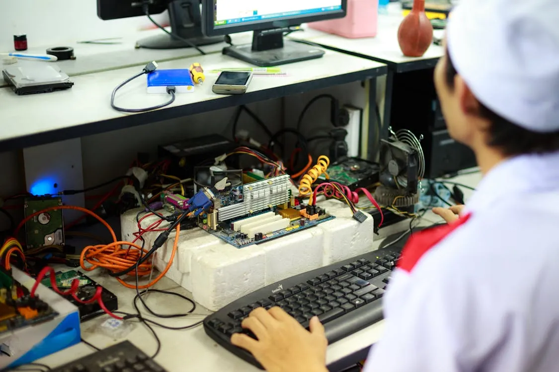 A man in a laboratory uniform working on computer components and wiring at a workstation, representing semi-legal hacking and hardware repair.