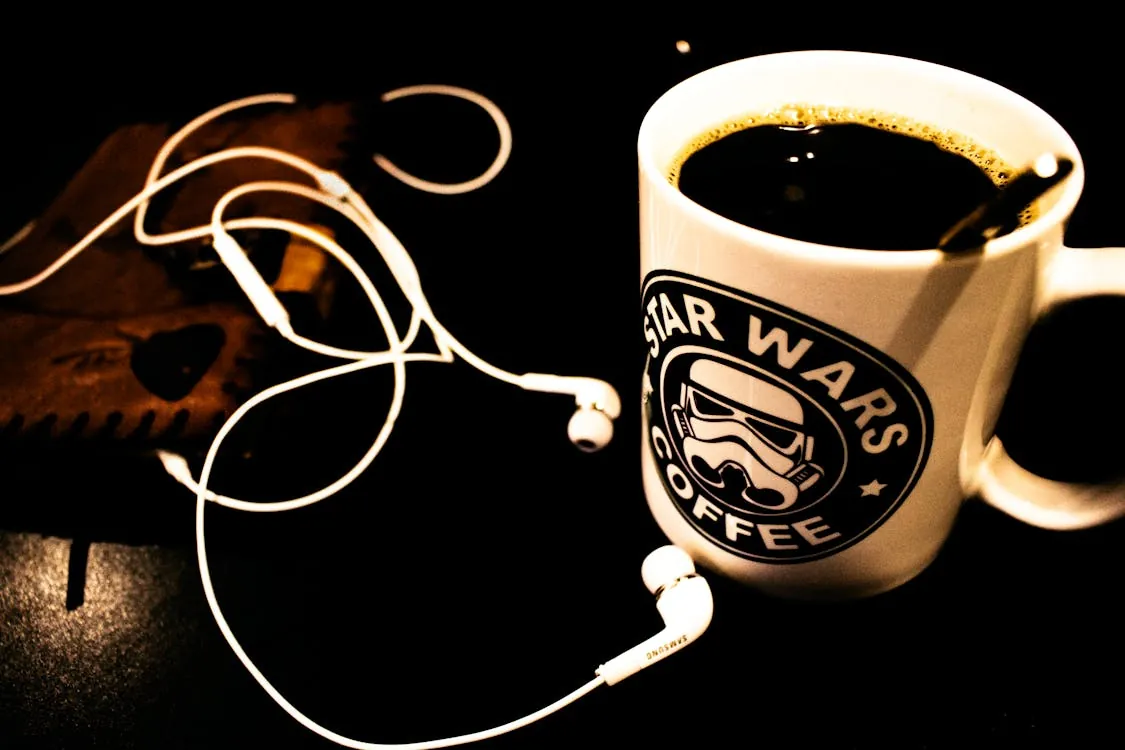 This photo shows a mug of black coffee with a "Star Wars Coffee" logo featuring a Stormtrooper helmet, placed on a dark surface next to a pair of white earbuds and a small leather wallet, suggesting a cozy morning or workspace setting ideal for a blog post about coffee, morning routines, or pop culture merchandise.