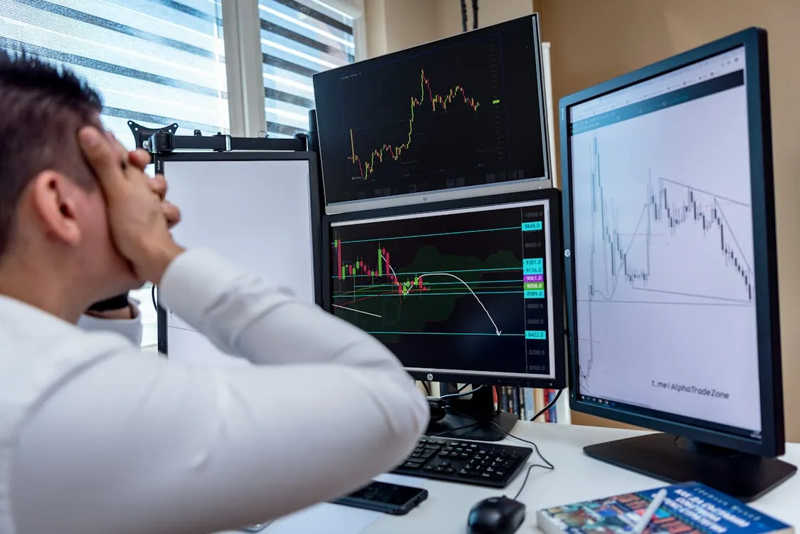 The photo shows a person sitting in front of multiple computer monitors displaying financial charts and stock or cryptocurrency data, with a frustrated or stressed expression, suggesting engagement in trading or market analysis. The setting appears to be an office or trading space, ideal for articles about financial markets, trading psychology, or investment strategies.