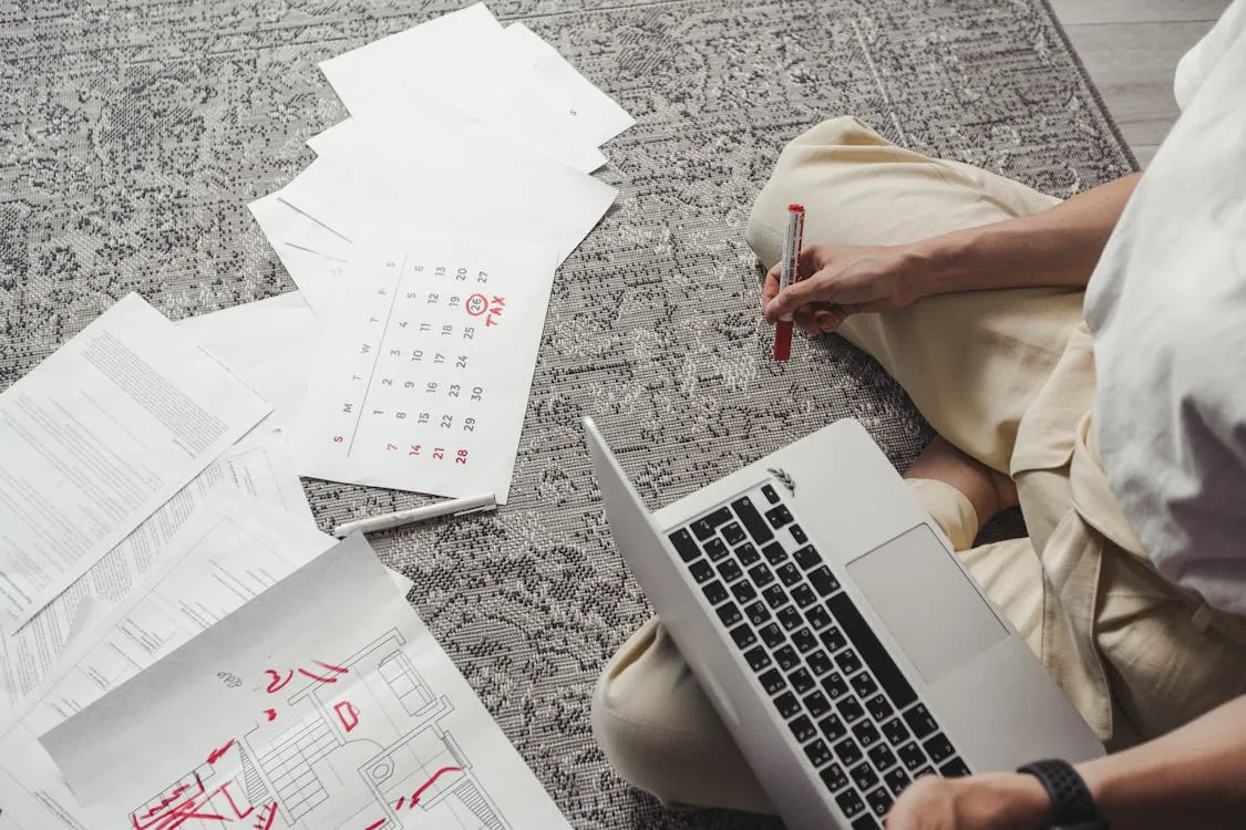 The image shows a person sitting cross-legged on a patterned rug, working with printed documents, a laptop, and a pen, suggesting they are engaged in planning or reviewing architectural or technical drawings. This setup indicates a focused work or study session, suitable for articles about remote work, planning, or design projects.