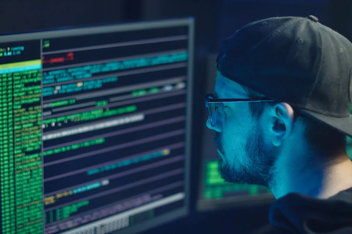 The image shows a man wearing glasses and a backward cap, focused on a computer screen displaying lines of code or data in various colors, suggesting an environment related to programming, cybersecurity, or hacking, with a dark background and multiple screens. This photo would be suitable for articles about coding, cybersecurity, hacking, or technology development.