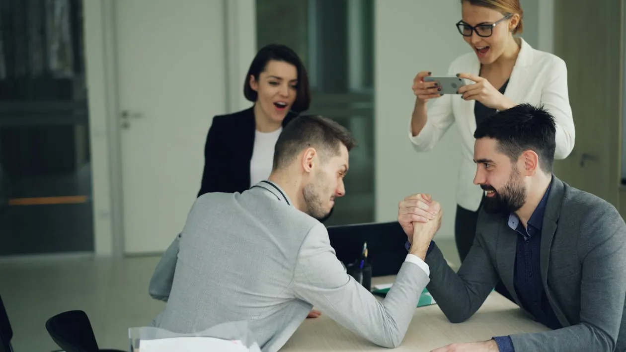 Two businessmen arm wrestling in an office with colleagues cheering them on, symbolizing competition in CI/CD pipeline tools.