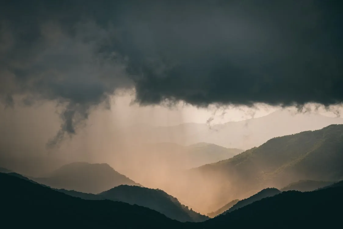 This photo depicts a moody, overcast mountain landscape with dark, ominous clouds overhead and a gentle cascade of rain or mist falling over the rolling hills in the background. It would be suitable for articles about weather, nature, outdoor adventures, or atmospheric scenery.