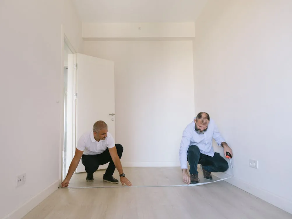 This photo shows two men measuring a section of the floor in a bright, minimally furnished room with white walls and light wood flooring, likely preparing for flooring installation or renovation. One man is crouched near the doorway, while the other is seated on the floor, both focused on the measuring tape, suggesting a focus on home improvement or interior decorating.