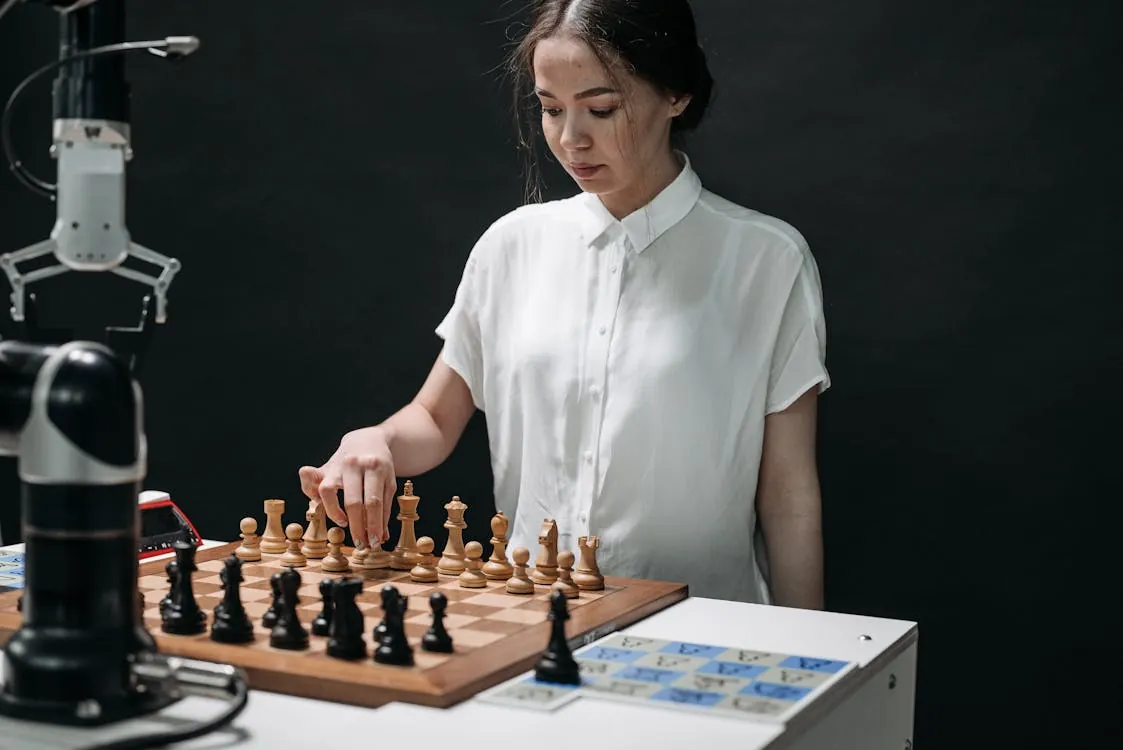 The image shows a young woman playing chess in a studio setting, with a dark background and professional equipment like a camera and lighting nearby, suggesting a photoshoot or recording session focused on chess or strategic thinking. It would suit an article about chess, mental games, or female players in competitive contexts.
