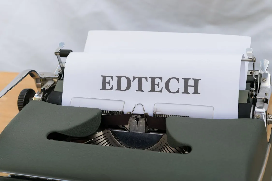 This image shows a vintage typewriter on a wooden surface with a piece of paper displaying the word "EDTECH" in bold, uppercase letters, set against a blurred, light-colored background, suggesting a focus on education technology.
