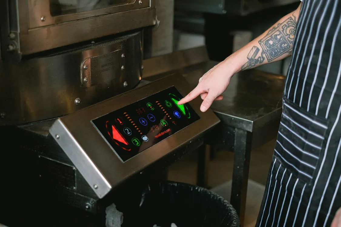 This photo shows a person in a black and white striped apron operating a modern, touchscreen commercial washing machine in a laundry or self-service laundry facility. The person is pointing at the control panel, which displays colorful digital icons and settings, highlighting the machine's advanced interface.