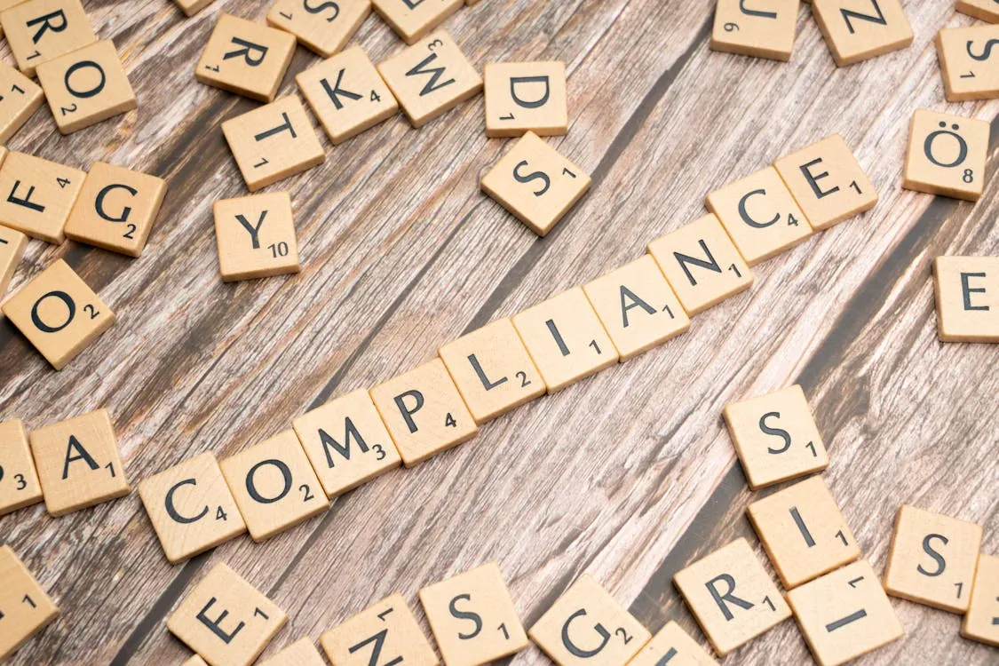 This photo shows a collection of Scrabble tiles arranged on a wooden surface, spelling out the word "COMPLIANCE," with scattered tiles nearby. It is suitable for an article discussing topics related to regulations, adherence, or corporate responsibility.