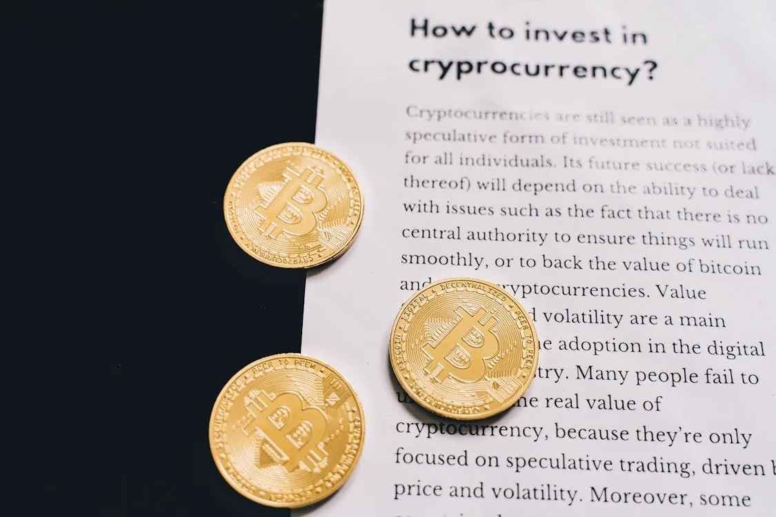 The image shows three gold-colored Bitcoin coins placed on a document titled "How to invest in cryptocurrency?" The coins are arranged on a white sheet of paper with text discussing cryptocurrency investment, suggesting a focus on digital currency education or financial advice.