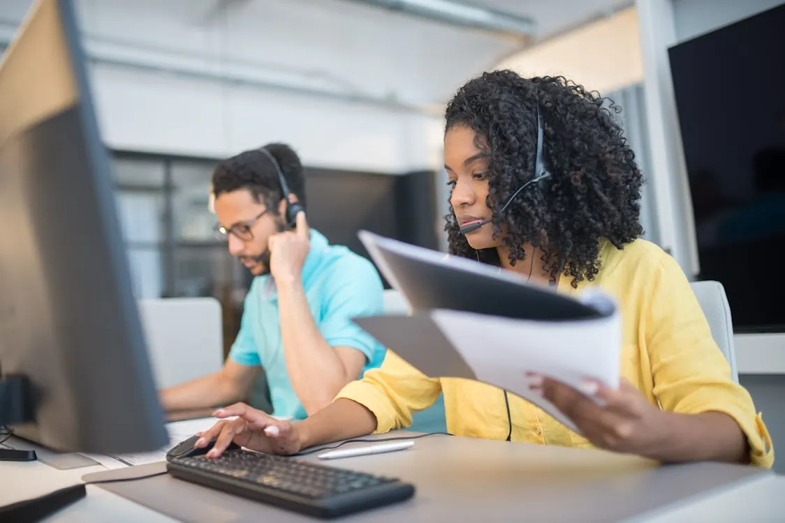 This image shows two customer service representatives working at their desks in an office, each wearing a headset and focusing on their computers, with the woman in the foreground reviewing printed documents. The setting appears professional, highlighting their roles in providing support or technical assistance.