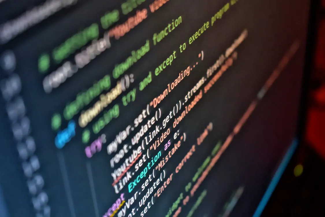 This photo shows a close-up of a computer screen displaying a colorful, syntax-highlighted code snippet, likely related to web development or programming, with text in multiple colors including green, yellow, purple, white, and red against a dark background. The image emphasizes the technical nature of coding, making it suitable for articles about software development, programming tutorials, or tech workflows.