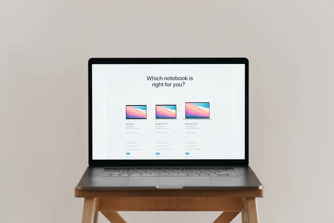 This image shows a laptop placed on a small wooden table, displaying a webpage asking "Which notebook is right for you?" with images and options comparing three different MacBook models. It would suit articles about selecting the right laptop or technology comparison guides.
