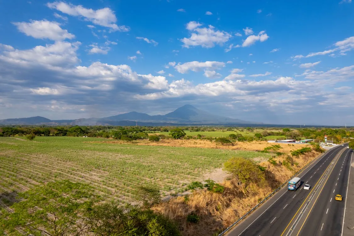 A photorealistic scene of a modern family with laptops and backpacks walking through a vibrant city street in El Salvador, with clear blue skies and lush green surroundings.