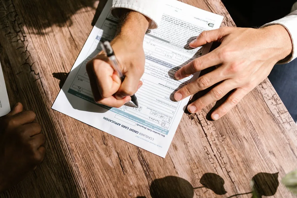 The image shows a person filling out a construction card application form at a wooden table, with another person nearby. It highlights the process of paperwork or documentation, suitable for articles on construction industry procedures, administrative tasks, or official forms.