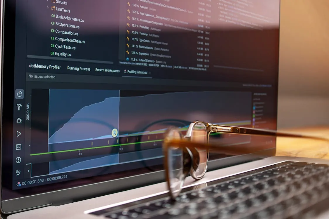 This photo shows a close-up of a laptop screen displaying code and performance profiling graphs, with a pair of eyeglasses resting on the keyboard, suggesting a focus on programming or software development in a professional or workspace setting.
