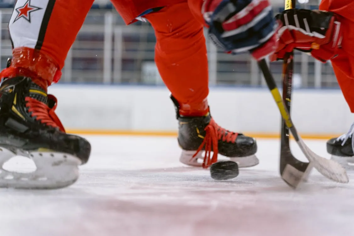 This photo shows a close-up of two ice hockey players' legs and equipment, with one player controlling the puck using their stick on an indoor rink. The focus is on their skates, hockey sticks, and the lower parts of their uniforms, highlighting the action and athletic gear involved in the game.