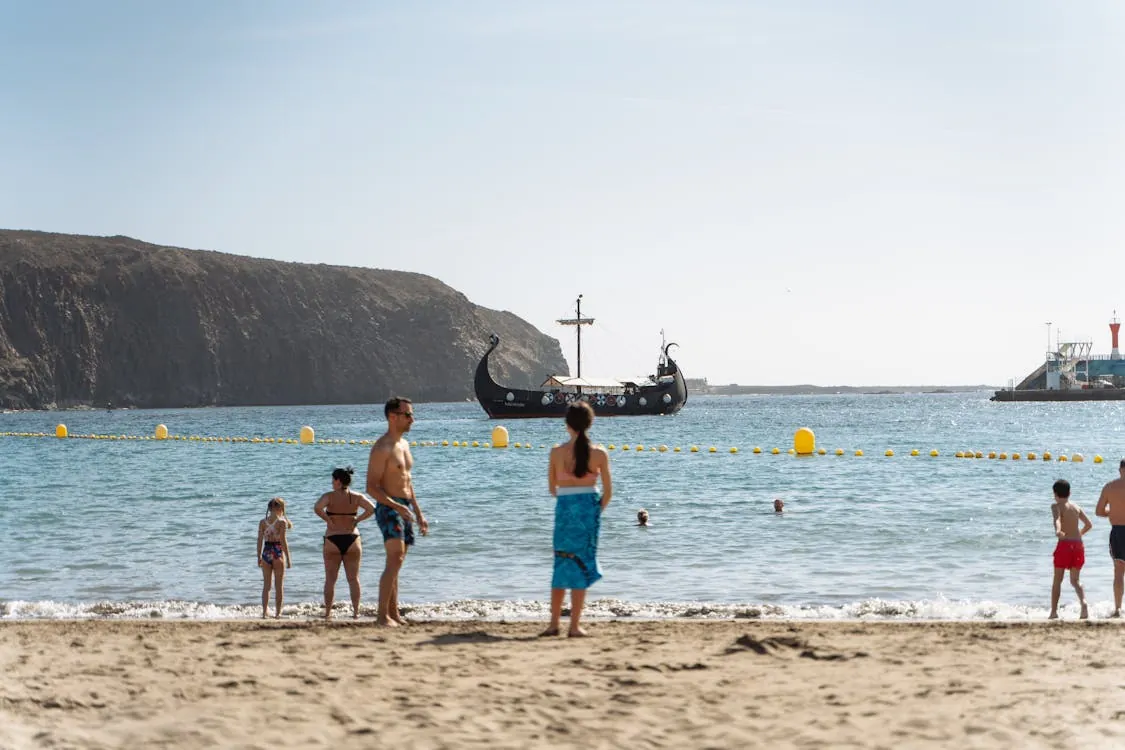 This photo shows a busy beach scene with families and individuals enjoying the water and sand under a clear sky; a Viking-style boat is anchored offshore, with a rocky coastline and lighthouse in the background, suggesting a scenic seaside location ideal for summer or vacation-related content.
