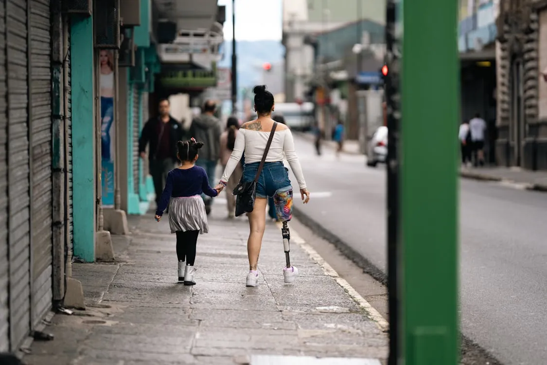 This photo shows a woman with a prosthetic leg holding hands with a young girl on a city sidewalk, with shops and pedestrians visible in the background. It captures a candid moment of daily life, highlighting themes of resilience, inclusivity, and urban environment.