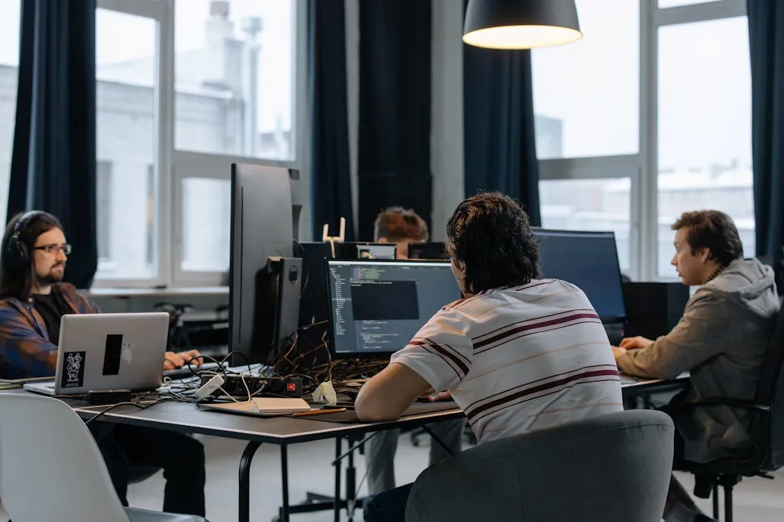 The photo shows a group of young adults working on computers in a modern, well-lit office setting, with multiple screens and coding visible on one monitor. The environment suggests a collaborative tech or software development workspace, suitable for articles about team programming, tech startups, or remote work culture.